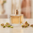 Un flacon de parfum carré vaporisant une brume, entouré de pistaches fraîches sur une surface en marbre.