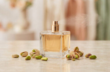 Un flacon de parfum carré vaporisant une brume, entouré de pistaches fraîches sur une surface en marbre.