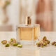Un flacon de parfum carré vaporisant une brume, entouré de pistaches fraîches sur une surface en marbre.