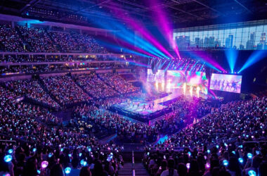 Vue panoramique d'un concert d'EXO avec une foule tenant des bâtons lumineux et une scène illuminée de néons.