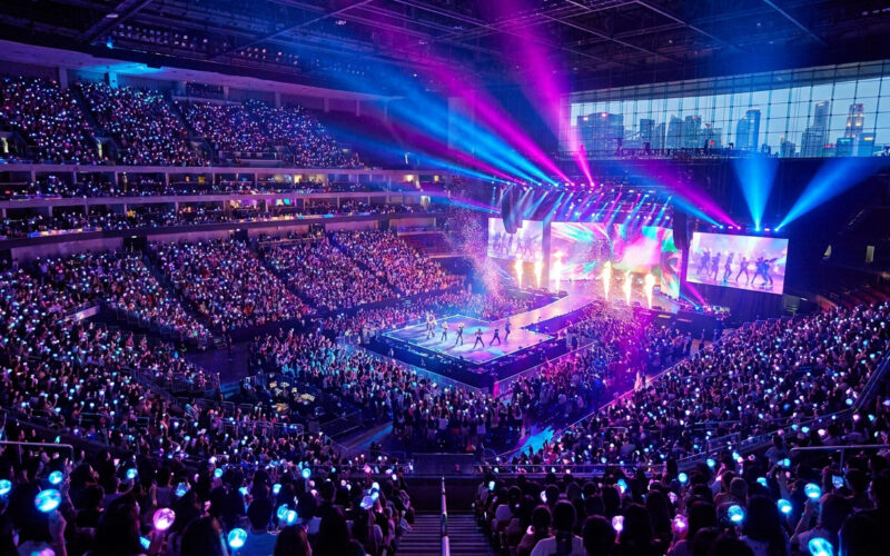 Vue panoramique d'un concert d'EXO avec une foule tenant des bâtons lumineux et une scène illuminée de néons.