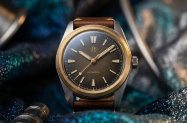 Montre automatique avec cadran marron, lunette dorée et bracelet en cuir, posée sur un tissu bleu brillant.