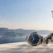 Une montre Zenith avec cadran bleu posée sur une terrasse surplombant la mer et des montagnes sous un ciel ensoleillé.